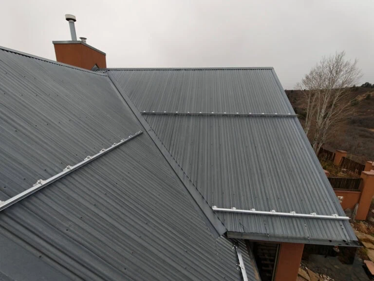 A close-up view of a metal roof with gray corrugated panels and snow guards, attached to a building under an overcast sky. A leafless tree and brown landscape are visible in the background.
