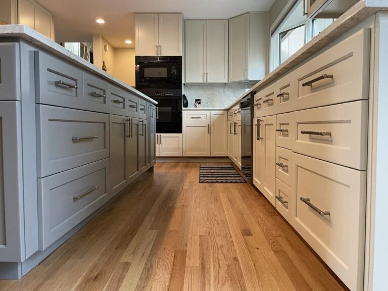 Modern kitchen with light wood floors, gray and white cabinets, stainless steel appliances, black oven and microwave, and a striped rug. The image is taken from a low angle between two counter islands.