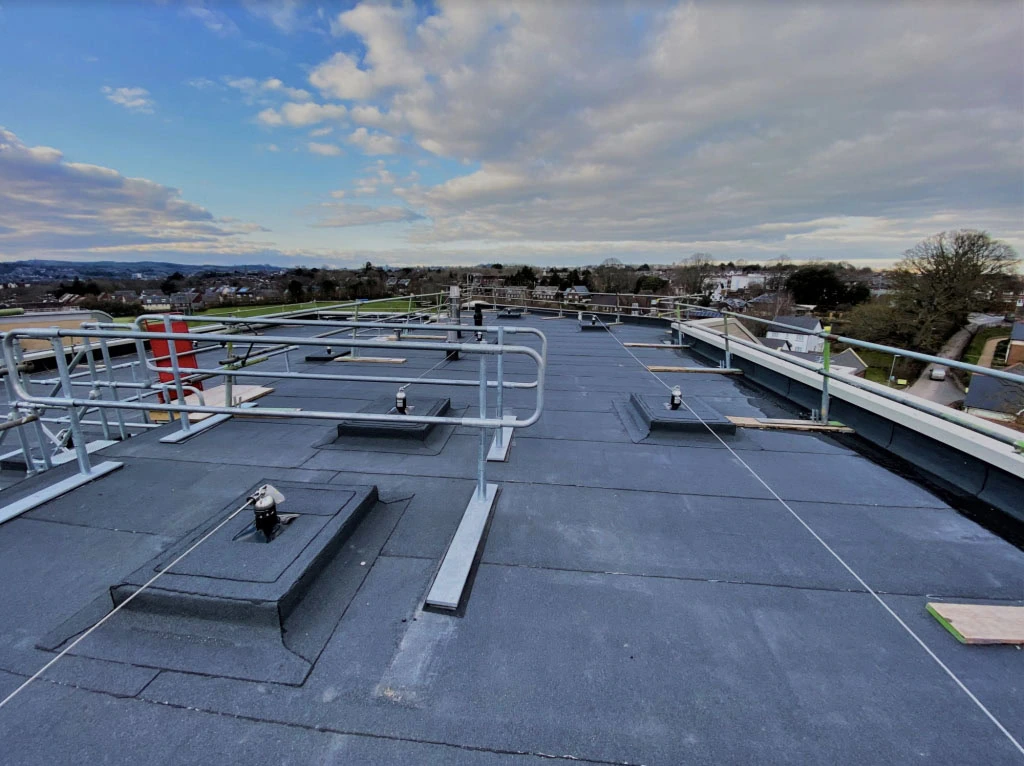Flat rooftop with safety railings, several metal anchors, and cables installed. The sky is partly cloudy, and a suburban area with trees and houses is visible in the background.