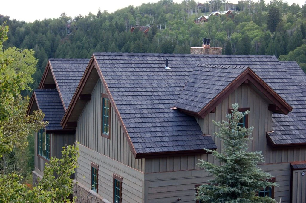 A house with a dark gray, shingle-style metal roof and taupe vertical siding is surrounded by evergreen trees and forested hills in the background.