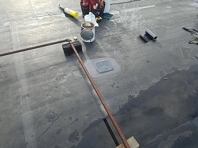 Roof repair tools and materials, including a rusted metal pipe, sealant, and a metal can, are placed on a flat, dark rooftop surface, showing patches and seams.