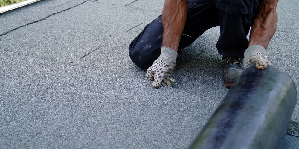 A person wearing gloves is installing a roll of roofing material on a flat roof, pressing it down to secure it in place. Only their arms and legs are visible.