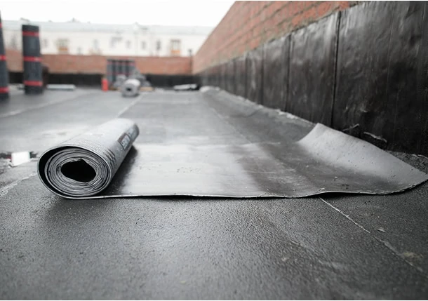 A roll of waterproofing membrane partially unrolled on a flat concrete rooftop, with brick walls on both sides and waterproofing material applied to the base of the walls in the background.