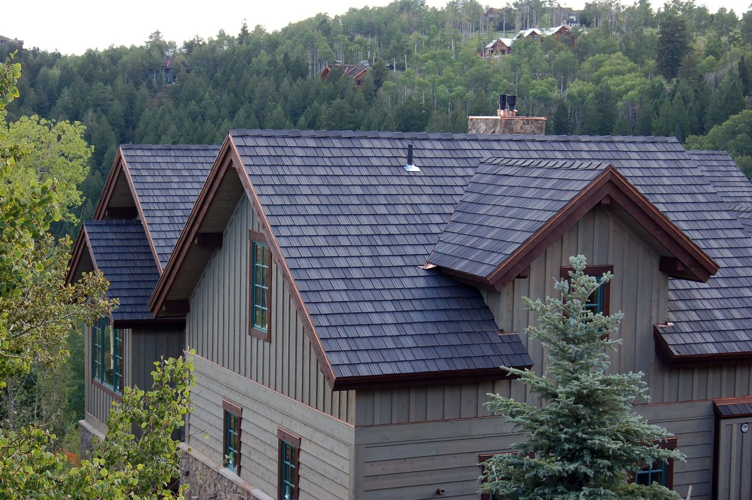 A large house with multiple peaked roofs covered in dark gray shingles, surrounded by green trees and forested hills in the background.