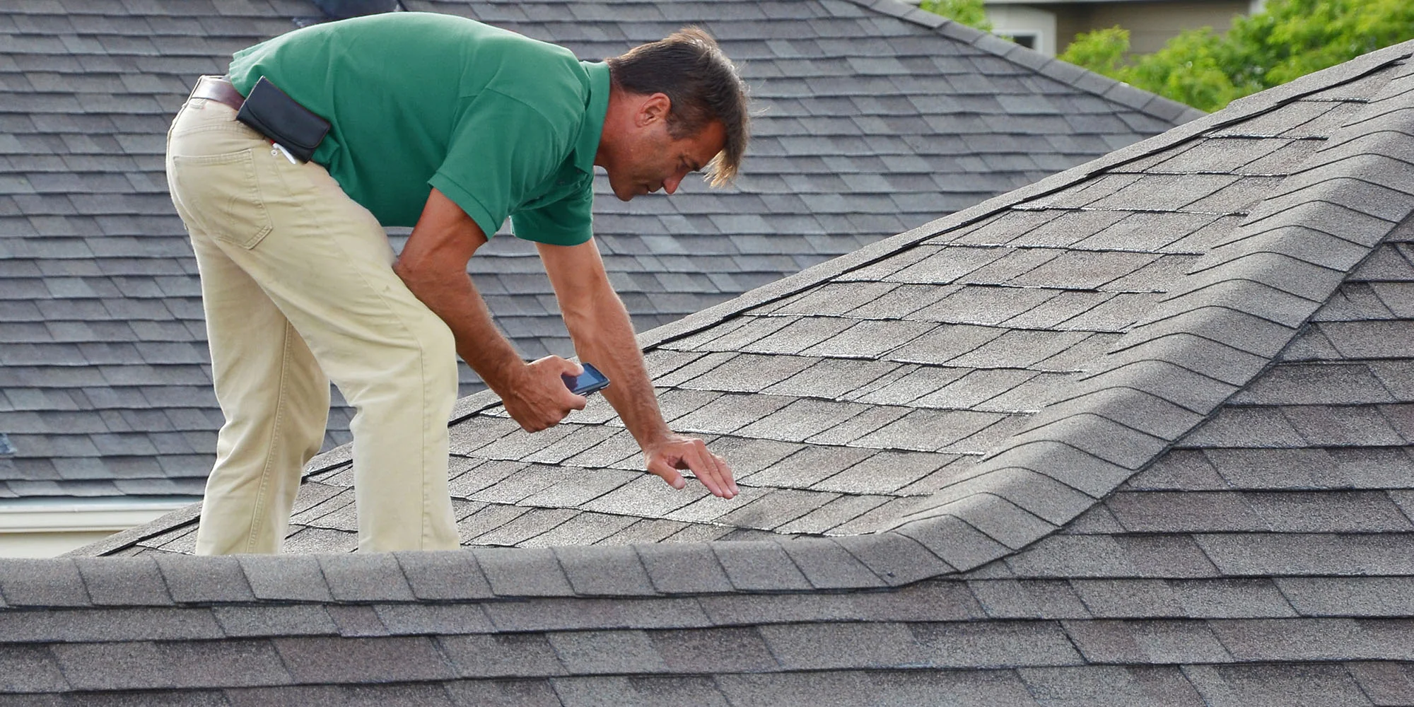 A man in a green shirt and khaki pants inspects the shingles on a gray, sloped roof, holding a mobile phone and closely examining the surface for damage.