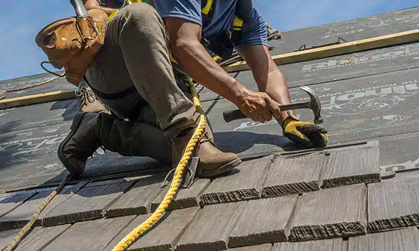 A construction worker wearing safety gear installs roofing shingles with a hammer, kneeling on a roof with protective underlayment and a safety harness attached.