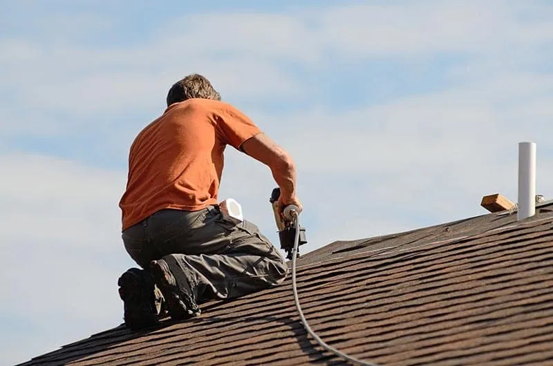 A person in an orange shirt and gray pants kneels on a sloped roof, using a tool to work on the shingles under a partly cloudy sky.