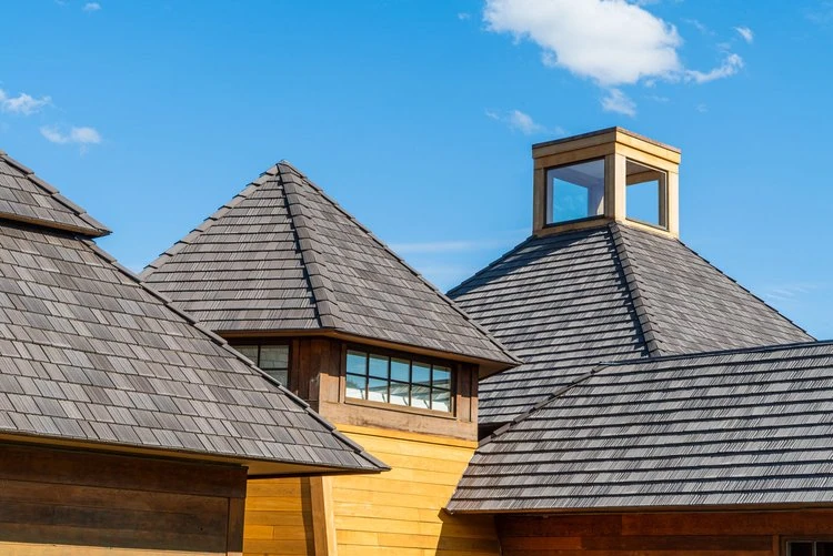 Close-up of rooftops with dark gray shingles and wooden siding under a blue sky. One roof features a cupola with windows, allowing light to enter. The architectural style appears modern and rustic.