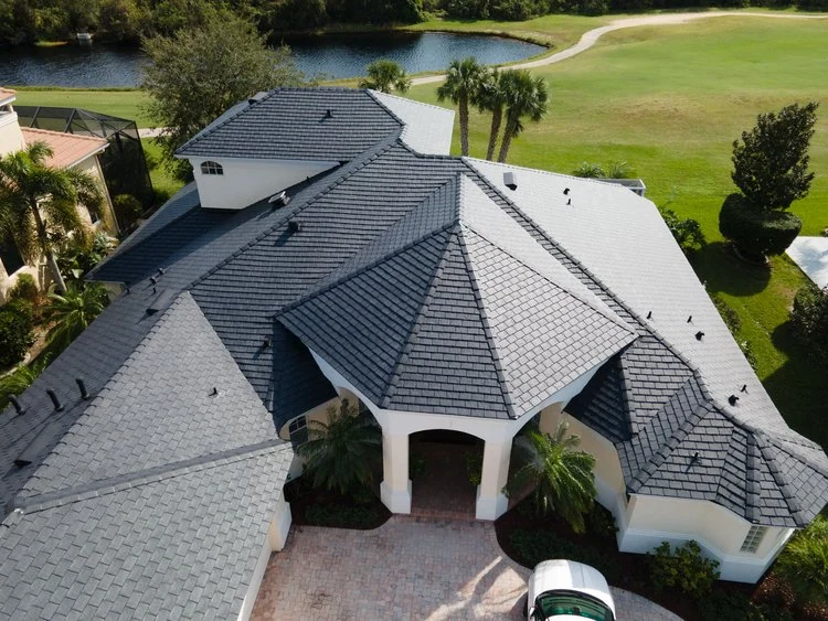 Aerial view of a large house with a gray, multi-gabled roof, surrounded by palm trees, a manicured lawn, a paved driveway, and a small lake with a golf course in the background.