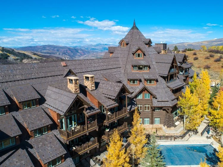 A large, multi-story wooden lodge with steep roofs sits among autumn trees and overlooks mountains under a blue sky. A pool is visible in the foreground.