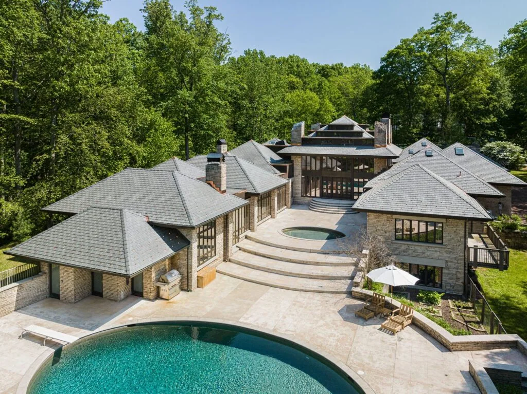 A large, modern stone house with multiple angled roof sections, situated in a wooded area. The backyard features a round pool, a hot tub, wide curved steps, and an outdoor patio with a table and umbrella.