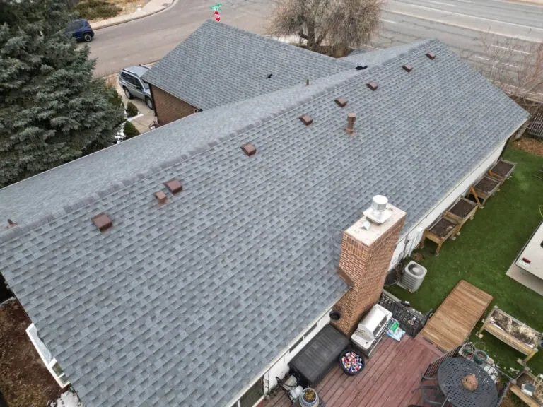 Aerial view of a house with a gray shingle roof, brown vents, a brick chimney, wooden deck, patio furniture, and a green lawn. A road and trees are visible in the background.