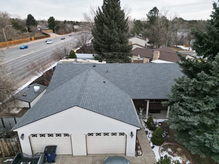 Aerial view of a house with a gray shingle roof, three-car garage, driveway, and trees. A road with light traffic runs alongside the property on a cloudy day.