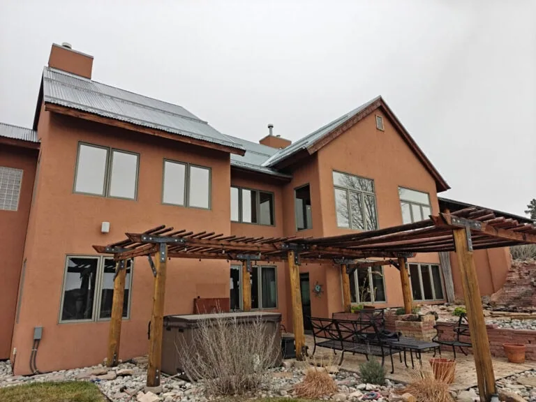 A modern two-story house with tan stucco exterior, large windows, a wooden pergola, patio furniture, and a hot tub, set in a landscaped backyard with rocks and sparse vegetation under a cloudy sky.