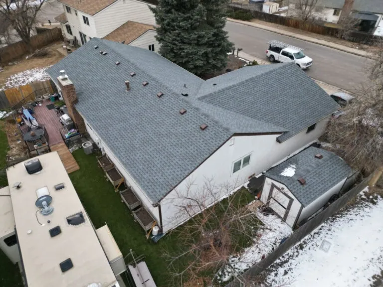 Aerial view of a gray-shingled house with a small shed, artificial grass yard, surrounding trees, some snow patches, and a white truck parked on the street.