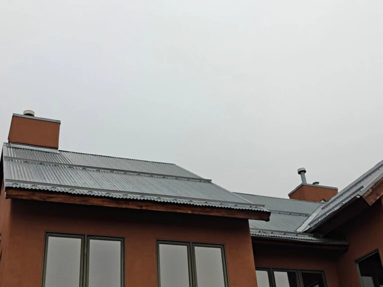 A modern building with brown walls, large windows, and a metal roof is shown under an overcast sky. The roof has two chimneys and angled sections.