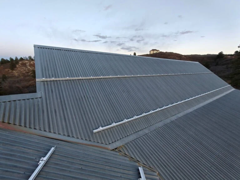 A large, corrugated metal roof with metal mounting rails installed on it, set against a backdrop of hills and a small structure in the distance under a cloudy sky.
