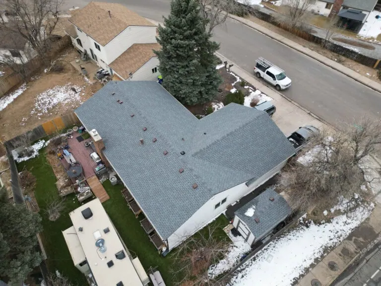 Aerial view of a house with a grey shingle roof, backyard deck, green lawn, and nearby vehicles. Light snow covers parts of the ground and rooftops.