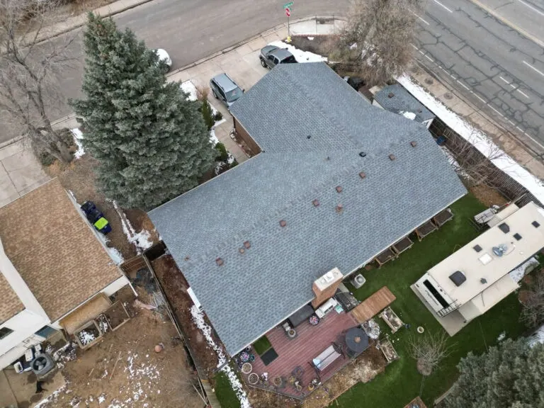 Aerial view of a house with a gray roof, backyard deck, patio furniture, nearby trees, grass, and an RV parked beside the yard. Snow patches are visible on the ground and rooftops.