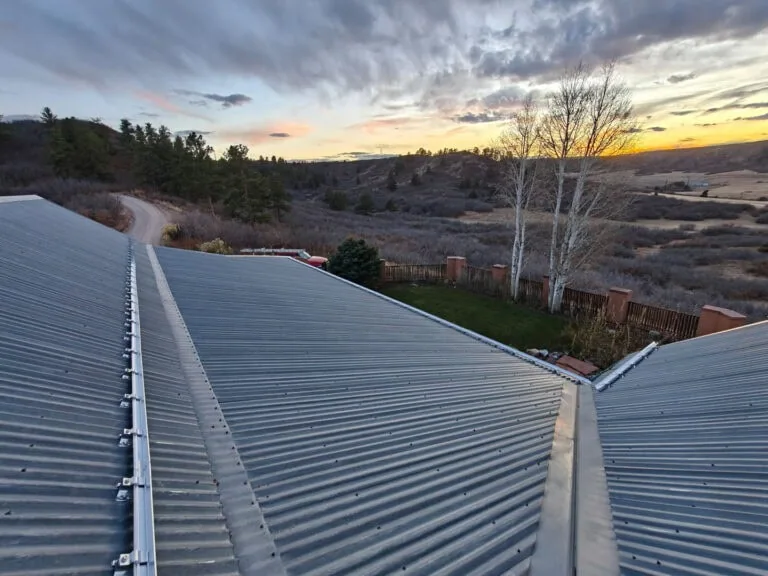View from a metal rooftop, overlooking a fenced yard, sparse trees, winding dirt road, and rolling hills at sunset with a colorful sky.