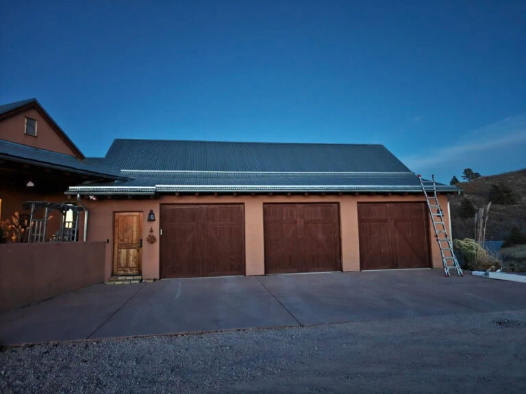 A house with three large wooden garage doors, a small front door, and a ladder leaning against the right side of the building at dusk. The sky is clear and blue.