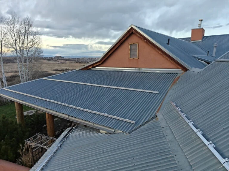 A close-up view of a metal roof with corrugated panels on a house, against a backdrop of trees, fields, and cloudy sky.