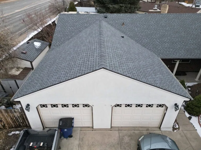 A gray shingled, double-car garage with white doors. Trash bins and a parked car are in the driveway, and patches of melting snow are visible around the garage and along the fence.