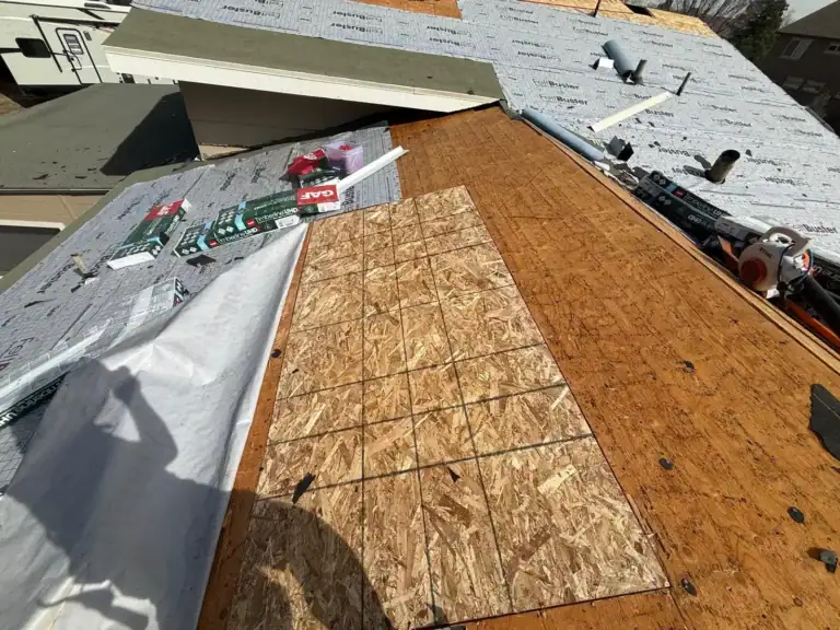 A partially repaired roof with a section of new plywood next to older wood, roofing materials, tools, and underlayment sheets spread out, with a persons shadow visible on the roof.