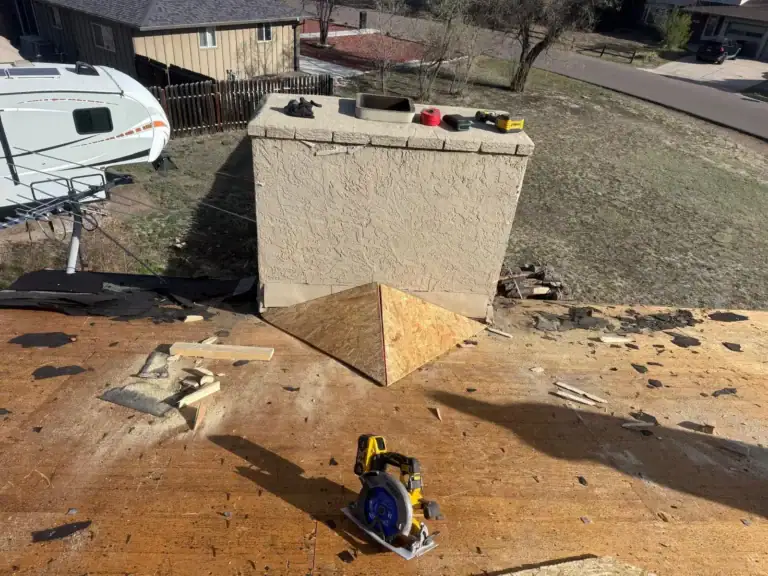 A rooftop under repair with tools, wood pieces, and a saw. A new wooden cricket structure is being built at the base of a chimney to divert water. Nearby are a house, an RV, and a yard.