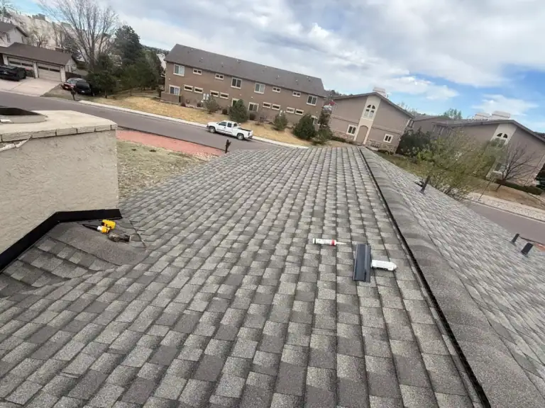 View from a rooftop covered with gray shingles, with tools resting on the roof. Houses, a white pickup truck, and trees are visible in the background under a partly cloudy sky.