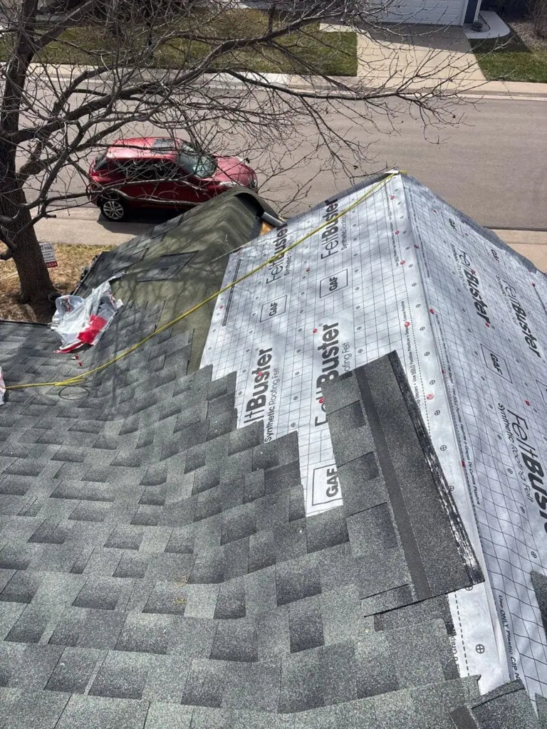 A view from a rooftop under construction, with partially installed shingles, roofing underlayment exposed, a tarp below, and a red car parked on the street beside a tree.