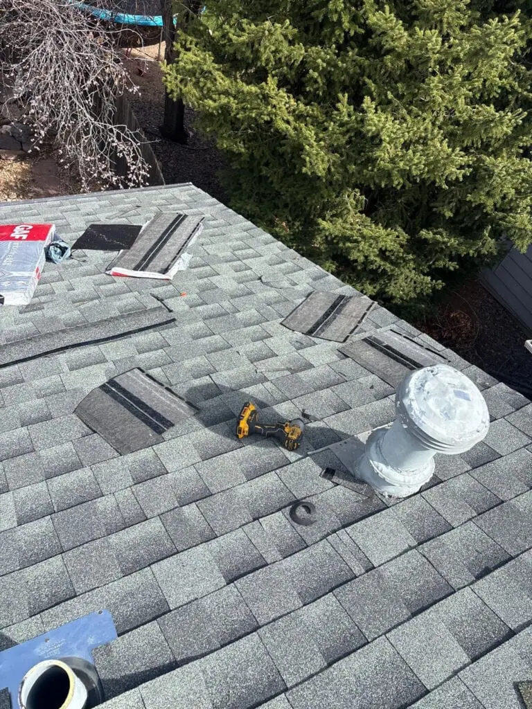 Gray shingles are being installed on a roof; some are loose or stacked. A power drill and vent pipe are visible. Trees and part of a yard are in the background.