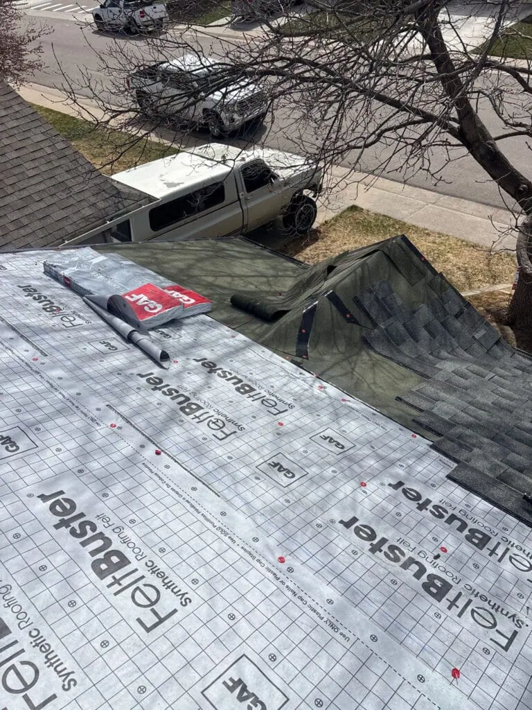 View from a roof under construction with synthetic roofing underlayment visible. Tools and packages are on the roof, with a tarp and a white van parked on the street below. Leafless tree branches are in the foreground.