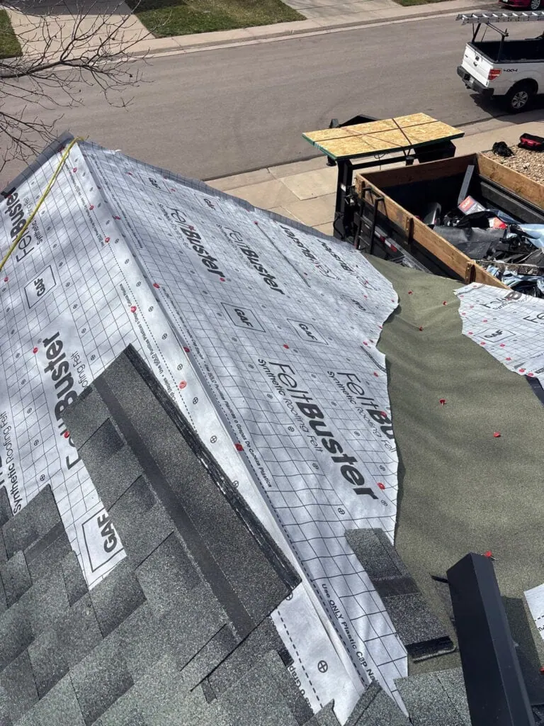 View of a house roof under construction, with roofing underlayment partially installed. Shingles are visible at the edges, and tools and materials are present on the ground and in a nearby trailer.