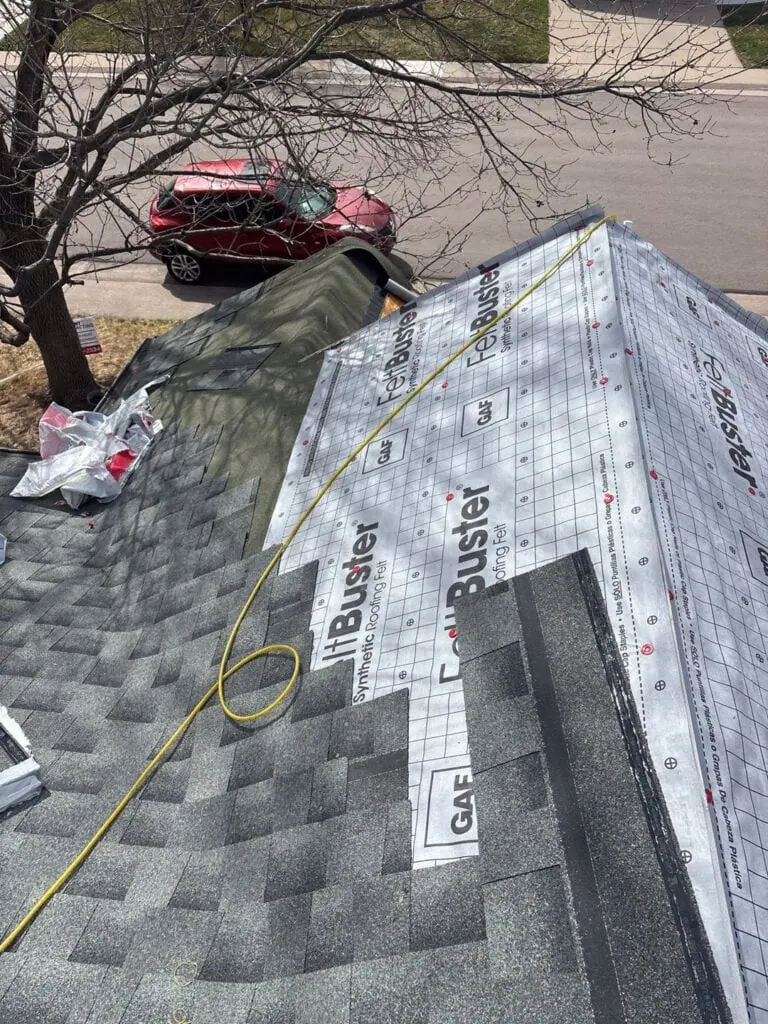 View of a house roof under construction, with shingles partially installed and synthetic underlayment visible. A yellow hose runs across the roof, and a red SUV is parked on the street below.