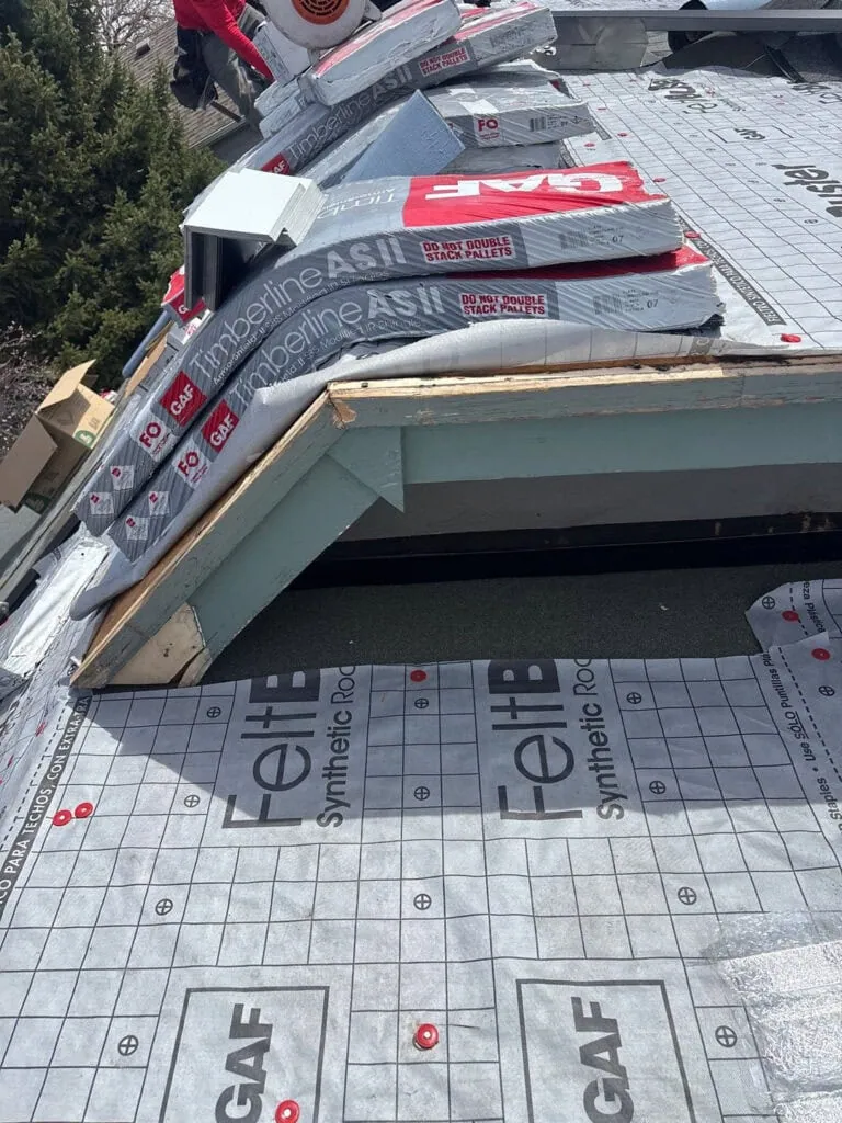 Roof under construction with GAF synthetic roofing underlayment and insulation boards stacked on top. Exposed wood and roofing materials are visible, along with tools and a worker’s arm in the background.