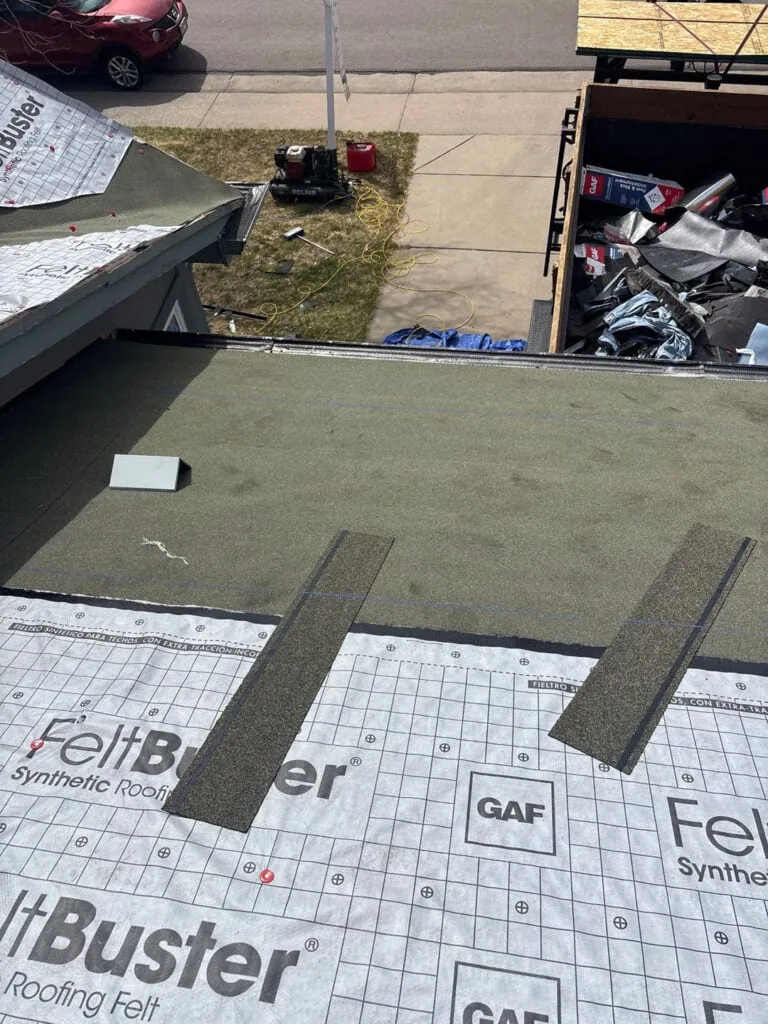 A view from a roof under construction shows roofing felt being installed, with tools and materials scattered below. Two asphalt shingles are placed on the roof, and a red car is parked beside the driveway.