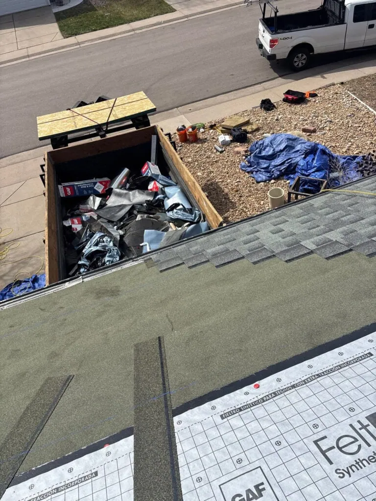 View from a roof under construction, showing roofing materials, debris in a dumpster, a truck parked by the curb, and tarps and tools spread on the ground near a landscaped yard.