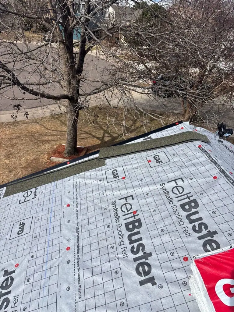 A rooftop covered with synthetic roofing felt is being worked on. A tree with bare branches is nearby, and a suburban street with houses and lawns is visible below.