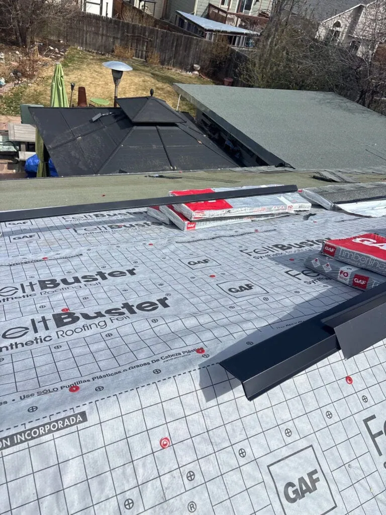 A rooftop with GAF FeltBuster synthetic roofing felt partially installed, roofing materials and shingle packs on top, and neighboring backyards visible in the background.