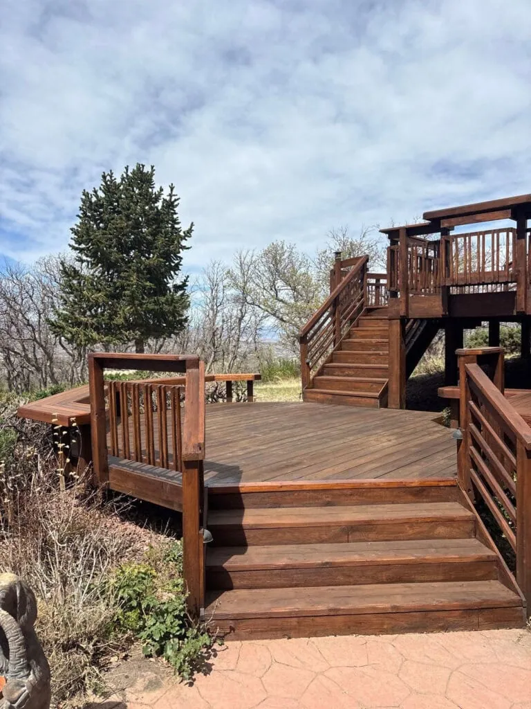 A large wooden deck with multiple levels and stairs sits outdoors, surrounded by bushes, bare trees, and a single evergreen. The sky is partly cloudy.