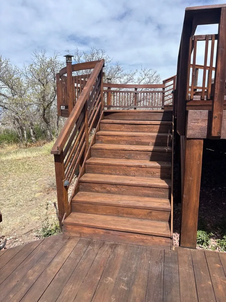 A set of wooden stairs with railings leads up to a deck surrounded by trees under a partly cloudy sky. The deck and stairs appear to be well-maintained and freshly stained.