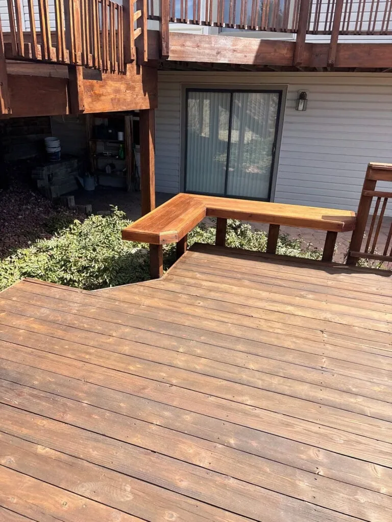 A wooden deck with built-in bench seating, attached to a house with white siding and a sliding glass door. The deck is elevated with railings, and greenery surrounds part of the deck.