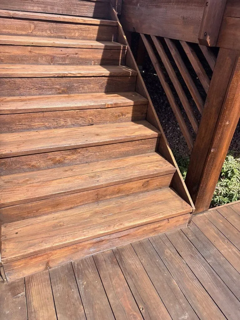 Wooden stairs leading up from a sunlit deck, with shadows cast across the steps and nearby railing. Small green plants are visible at the base.