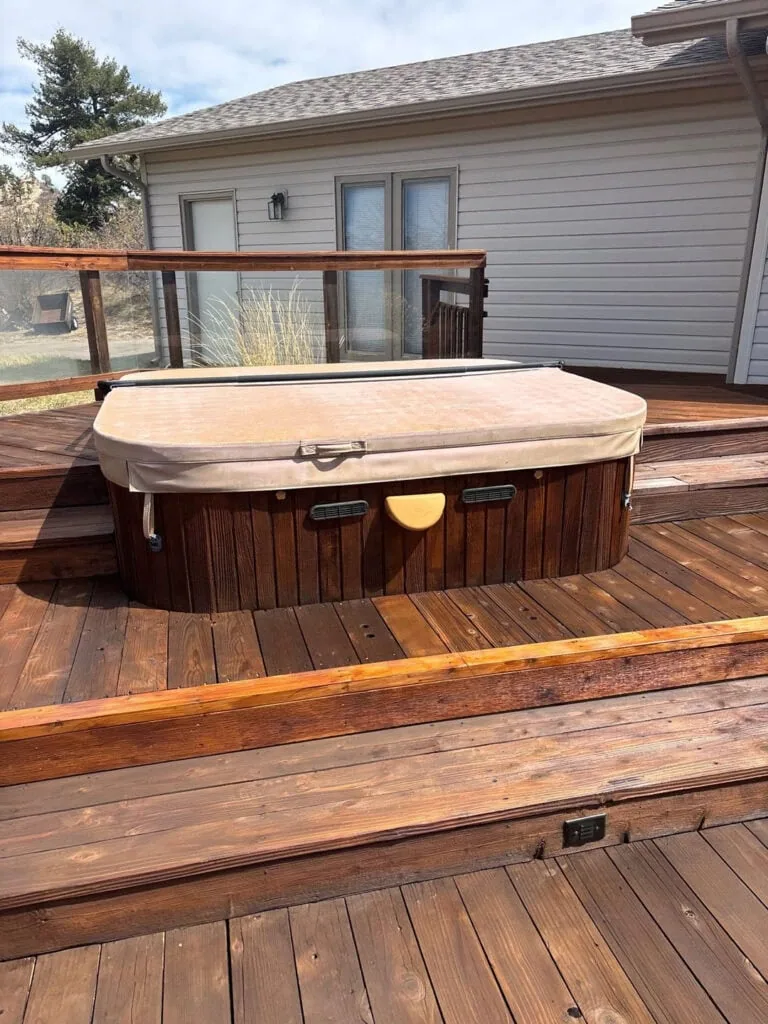 A closed hot tub with a tan cover sits on a wooden deck outside a house with beige siding and glass railing panels. The sky is partly cloudy.