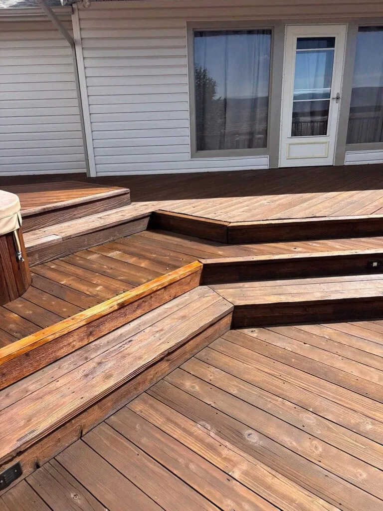 Wide wooden deck with multiple steps, a white house in the background, and a hot tub partially visible on the left. The deck boards have a warm, natural finish, and the steps create an angled geometric pattern.