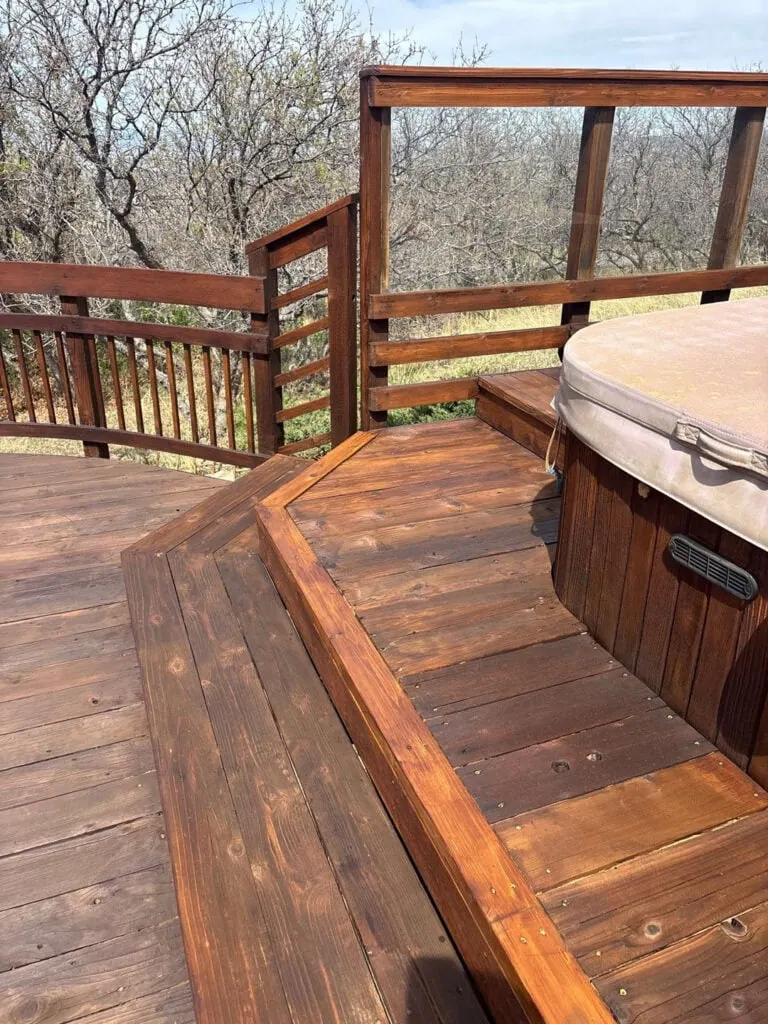 Wooden deck with steps leading to a circular hot tub. The deck has railings, and the background features leafless trees and a cloudy sky.