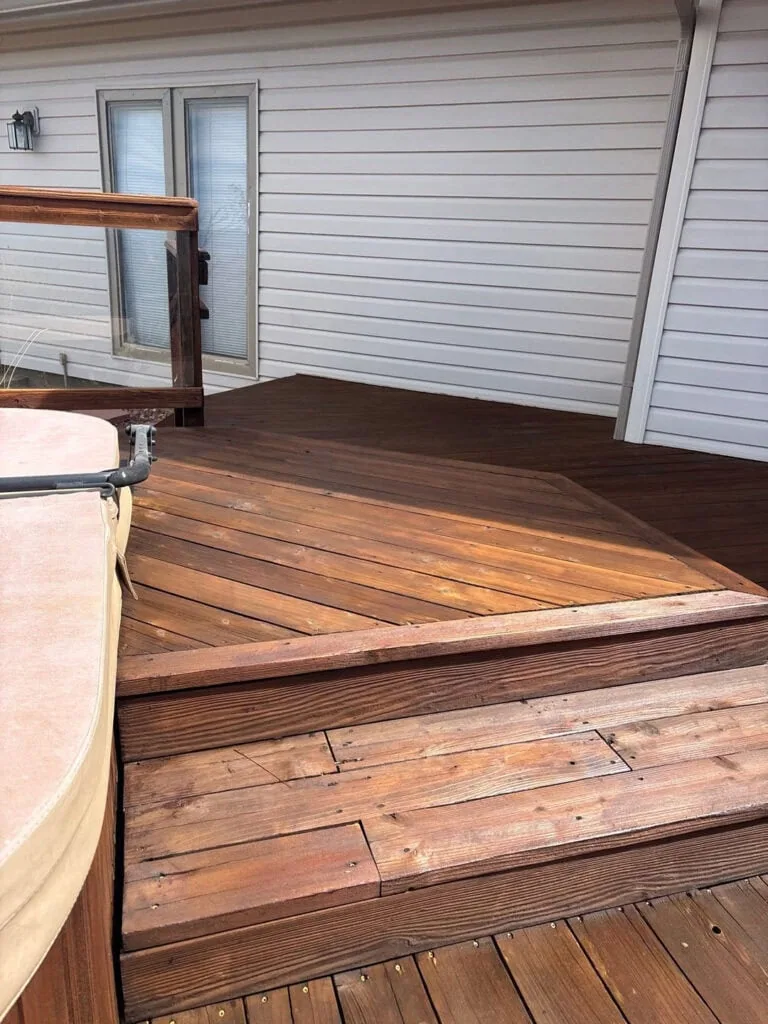 A wooden deck with steps leads to a hot tub on the left. The deck is attached to a white house with glass double doors. A railing is visible on the left side of the deck.