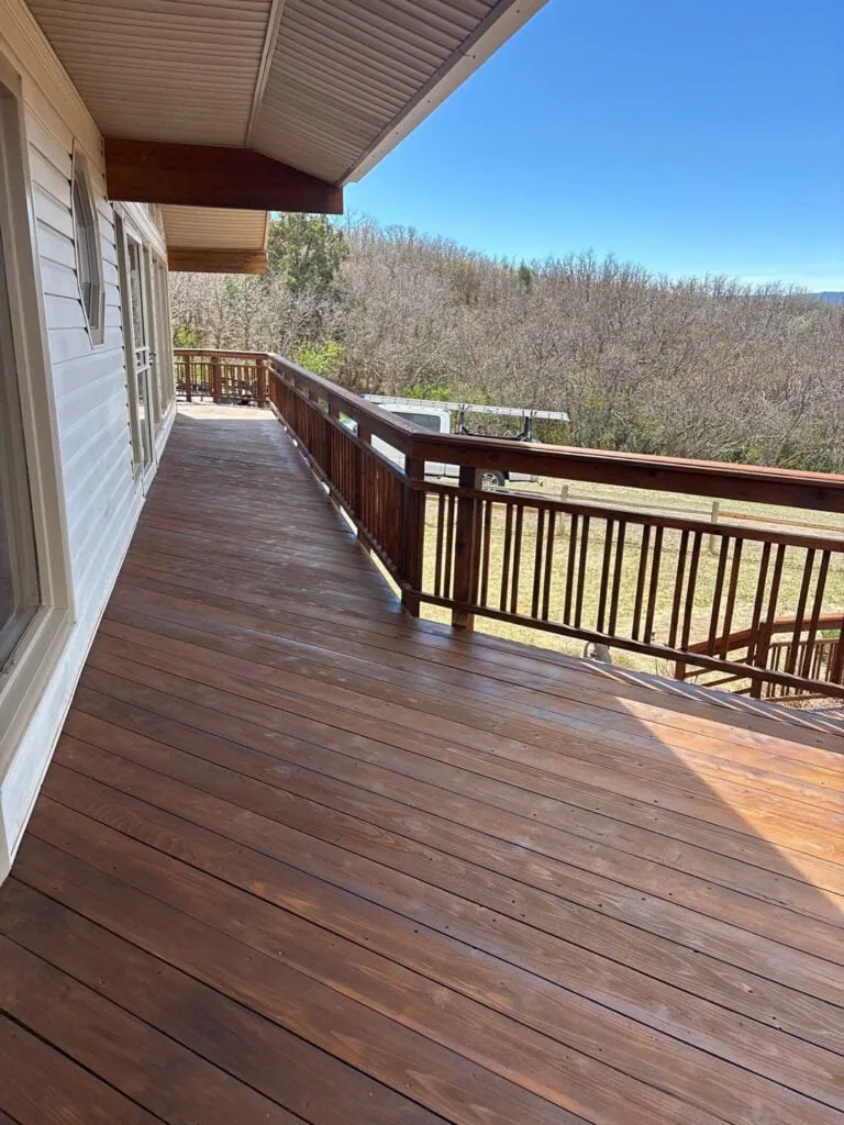 A spacious wooden deck with railings wraps around a house, overlooking a grassy yard and leafless trees under a clear blue sky.