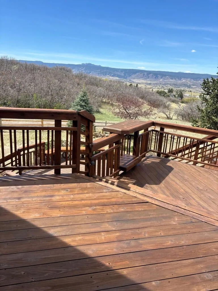 A wooden deck with stairs overlooks a grassy yard, trees, and distant mountains under a clear blue sky.
